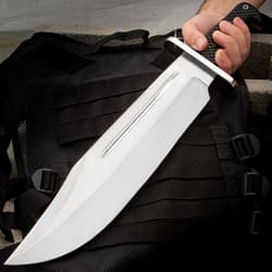 Large shining silver bowie knife being held at a downwards left angle on a background of grey concrete and a black tacticle bag.