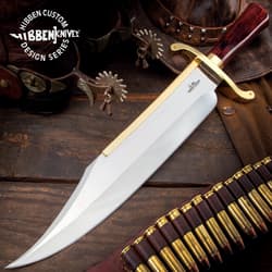 Large silver bowie knife with gold accents and a polished wood handle on a aged wood background with western props.