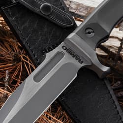 The Rambo Last Blood Bowie Knife rests on a black sheath. It features a satin-finish blade with “RAMBO” engraved near the hilt. The textured handle is secured by a polished screw.