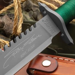 The Rambo I Signature Edition Knife shows an engraved signature and edition number. It features a sharp serrated spine, green cord-wrapped handle, and a partial view of the brown leather sheath.