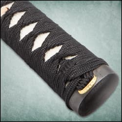 Close view of the brass menuki on the white ray skin handle with black braided cord.