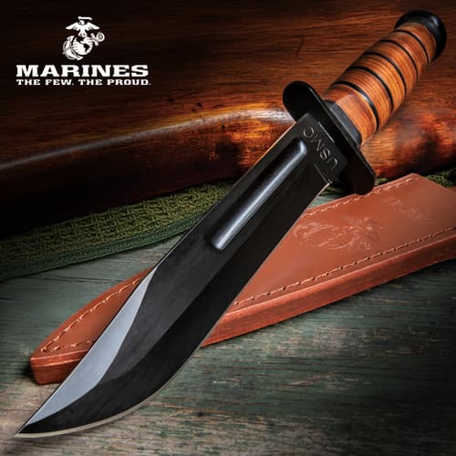 Fixed blade knife with stacked leather handle and large "USMC" engraving on a green antique wood background with a leather sheath and wooden gun handle.