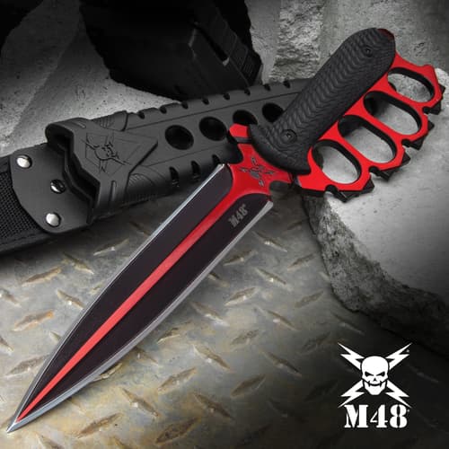 Double-edged trench knife with brass knuckle style handle with black oxide coating on a background of metal and concrete.