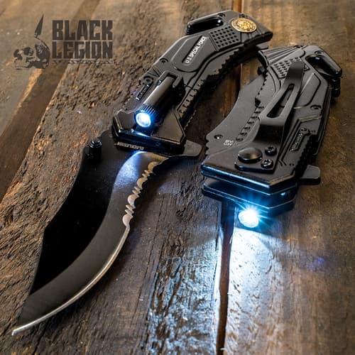 Black steel pocket knife with serated edge with illuminated flash attached on top, with identical closed knife to its right showing black pocket clip and seatbelt cutter on wooden background.