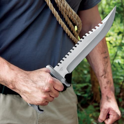 A hand is shown holding the knife by its black pakkawood handle with the sawback stainless steel blade facing upward.