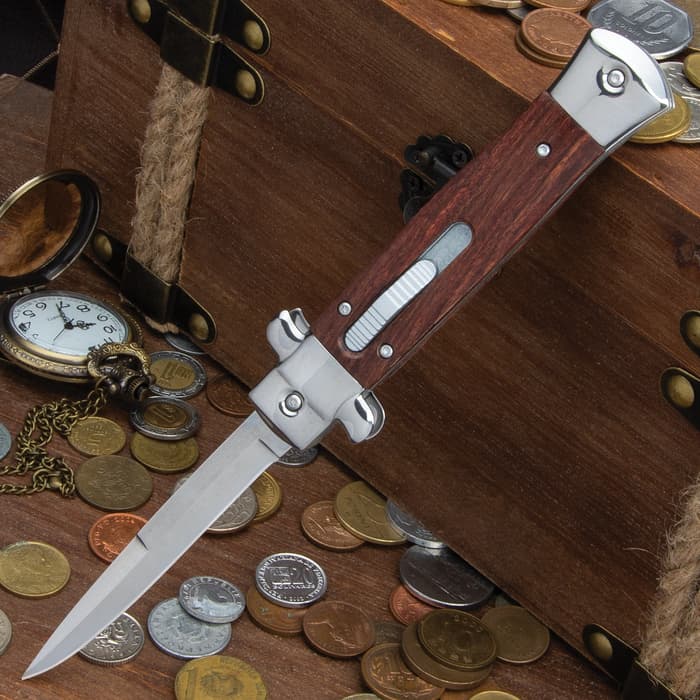 Wood handle stiletto knife with slide trigger and mirror polished accents on a background of wood and coins. 
