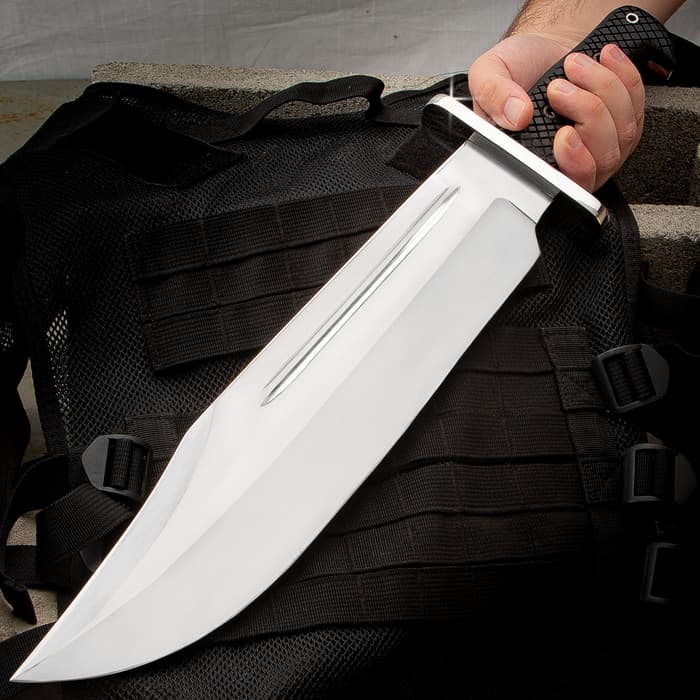 Large shining silver bowie knife being held at a downwards left angle on a background of grey concrete and a black tacticle bag.
