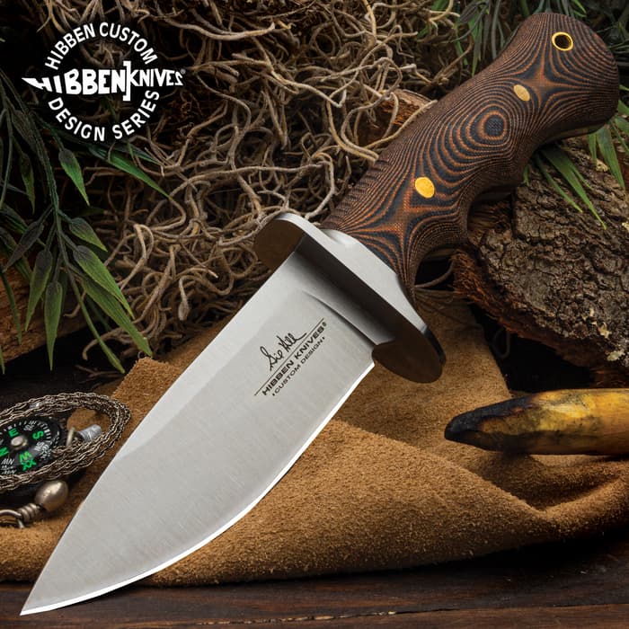 9 5/8" stainless steel fixed blade knife with G10 curved handle and brass pins on a background of moss, greenery, wood, and seude.
