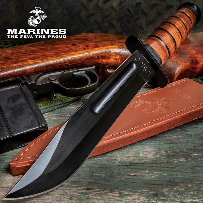 Fixed blade knife with stacked leather handle and large "USMC" engraving on a green antique wood background with a leather sheath and wooden gun handle.
