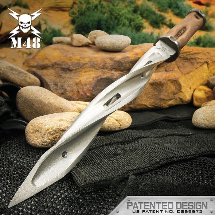 The M48 Battle Scarred Series Tan Cyclone has a 8” cast stainless steel blade with three spiraling cutting edges, shown leaned against rocks.