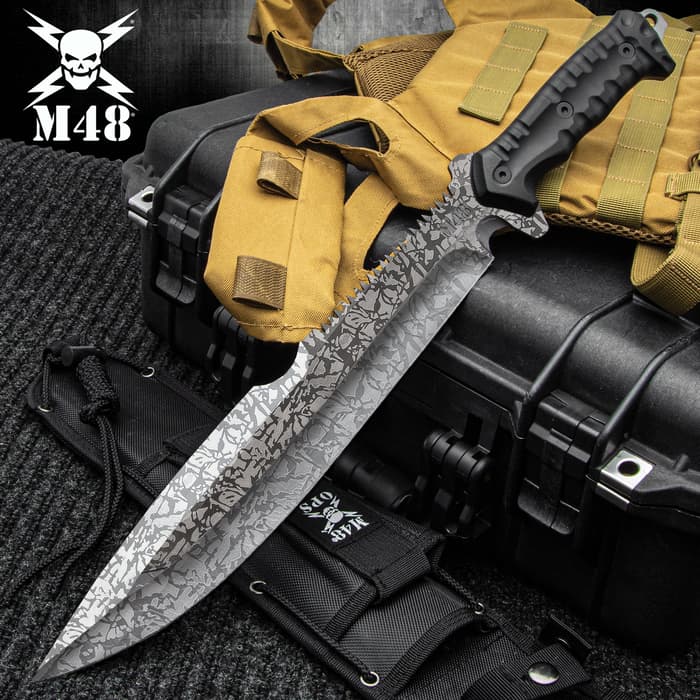 Combat machete with spiderweb titanium electroplated finish and a partial sawback spine on a background of black and tan utility gear. 
