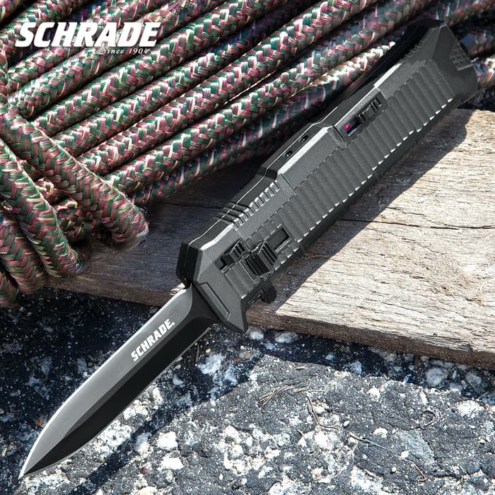 Sunlight reflects off of a satin black assisted opening pocket knife with a concert, wood, and rope background. Top left corner "Schrade."
