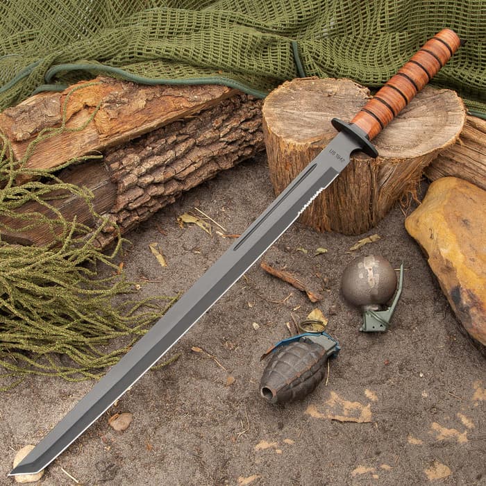1942 U.S. Combat Sword has a 19 3/4” black-coated AUS-6 stainless steel blade and a genuine stacked leather handle, shown laid against a stump.