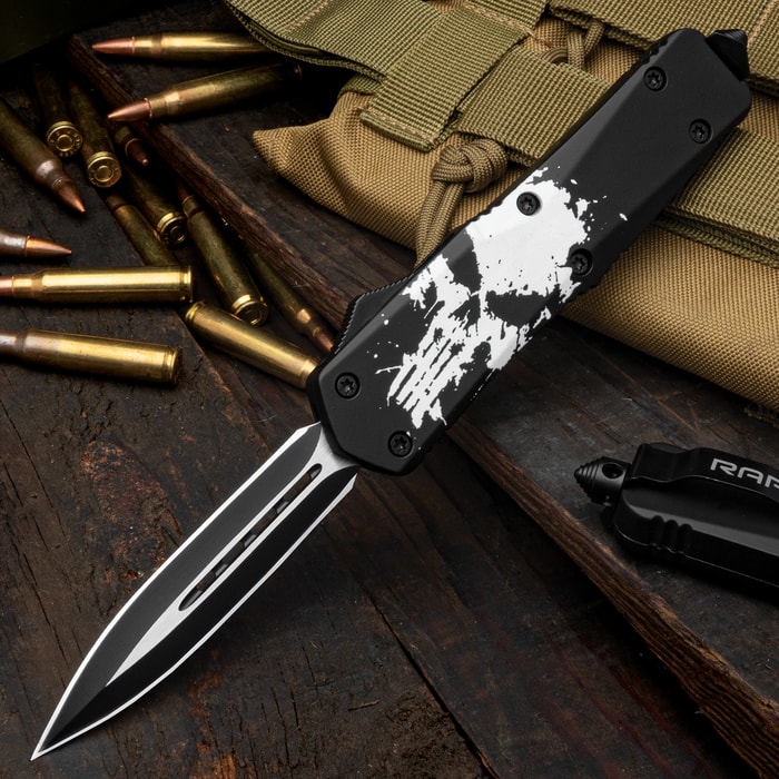 Extended OTF pocket knife with a dual toned matte black and silver dagger blade and black handle featuring a large distressed white skull print on a background of wood with bullets.