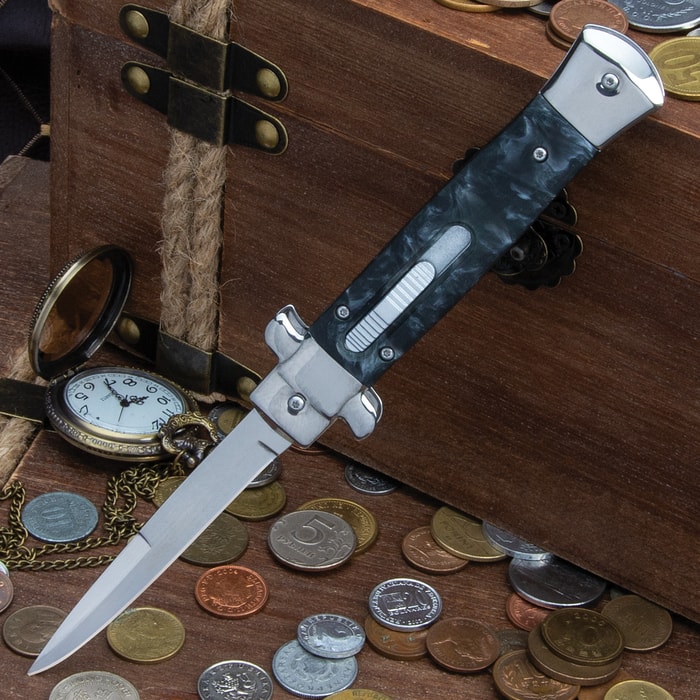 Automatic OTF Knife shown with black marbleized handle with blade deployed leaned against a wooden chest with coins.