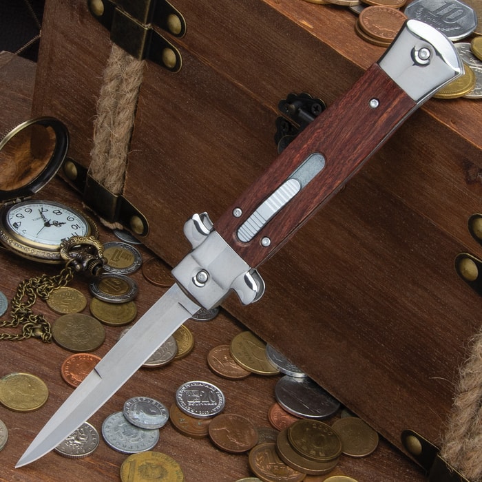 Automatic stiletto knife with wooden handle shown with blade deployed, leaned against a wooden chest with coins.