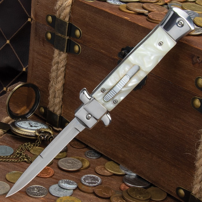 Stiletto automatic knife shown with blade deployed from the faux pearl handle, set against a wooden box with coins.