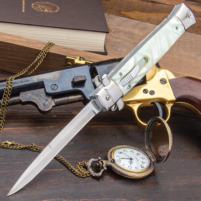 Large Automatic OTF Pearl Stiletto Knife with stainless steel blade and faux pearl handle, shown laid against a firearm and pocket watch.