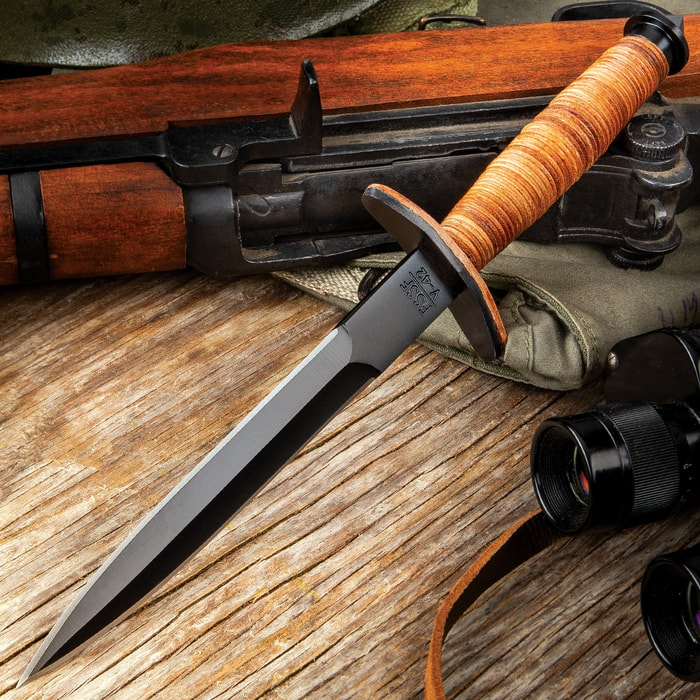 Stiletto dagger with a black blade finish and stacked leather handle on a background of wood and tactical gear.
