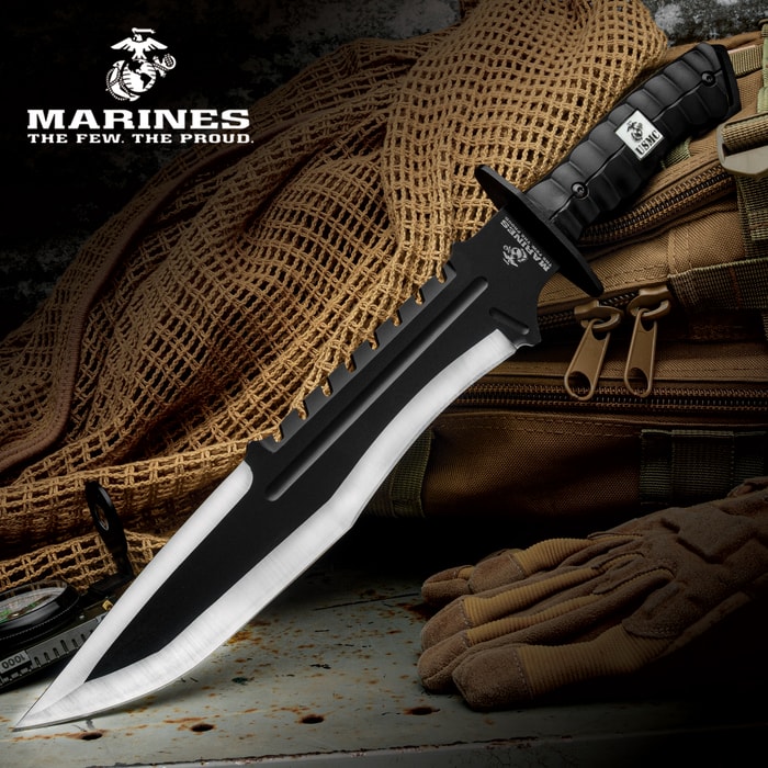The USMC Bulldog Bowie Knife with two-toned stainless steel blade and black textured handle shown beneath the Marines logo, laid on a tactical backpack.
