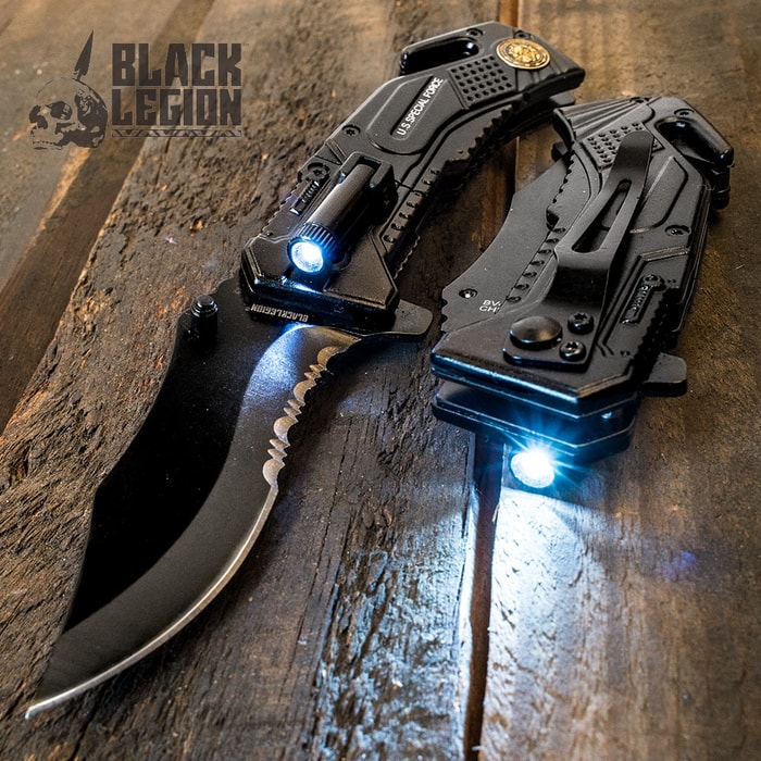 Black steel pocket knife with serated edge with illuminated flash attached on top, with identical closed knife to its right showing black pocket clip and seatbelt cutter on wooden background.
