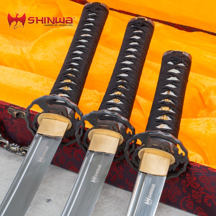 Three Shinwa samurai swords with black cord wrapped handles laid atop an open red display box. 
