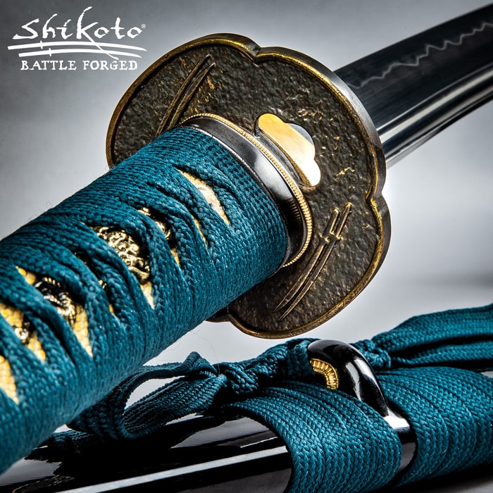 Katana handle wrapped in a teal cord and ray skin with a brass tsuba etched with double swords lying on a black scabbard
