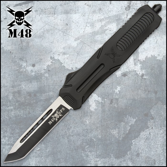 Matte black OTF tanto knife with a glass breaking pommel featuring a "M48" logo on both the blade and the handle on a background of tactical gear.