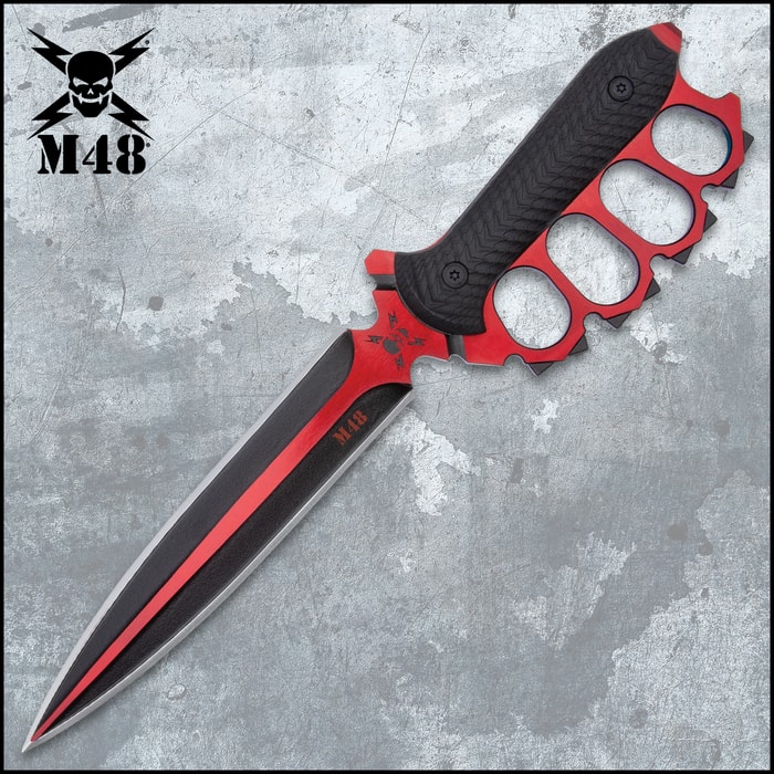 Double-edged trench knife with brass knuckle style handle with black oxide coating on a background of metal and concrete.